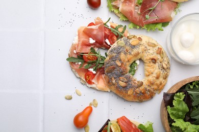 Tasty bagel with cured ham, cheese, tomatoes and arugula on white tiled table, top view. Space for text