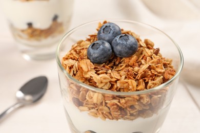 Photo of Glass of tasty yogurt with muesli and blueberries served on white table, closeup