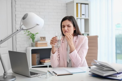 Young woman taking pill against headaches while sitting at table in office