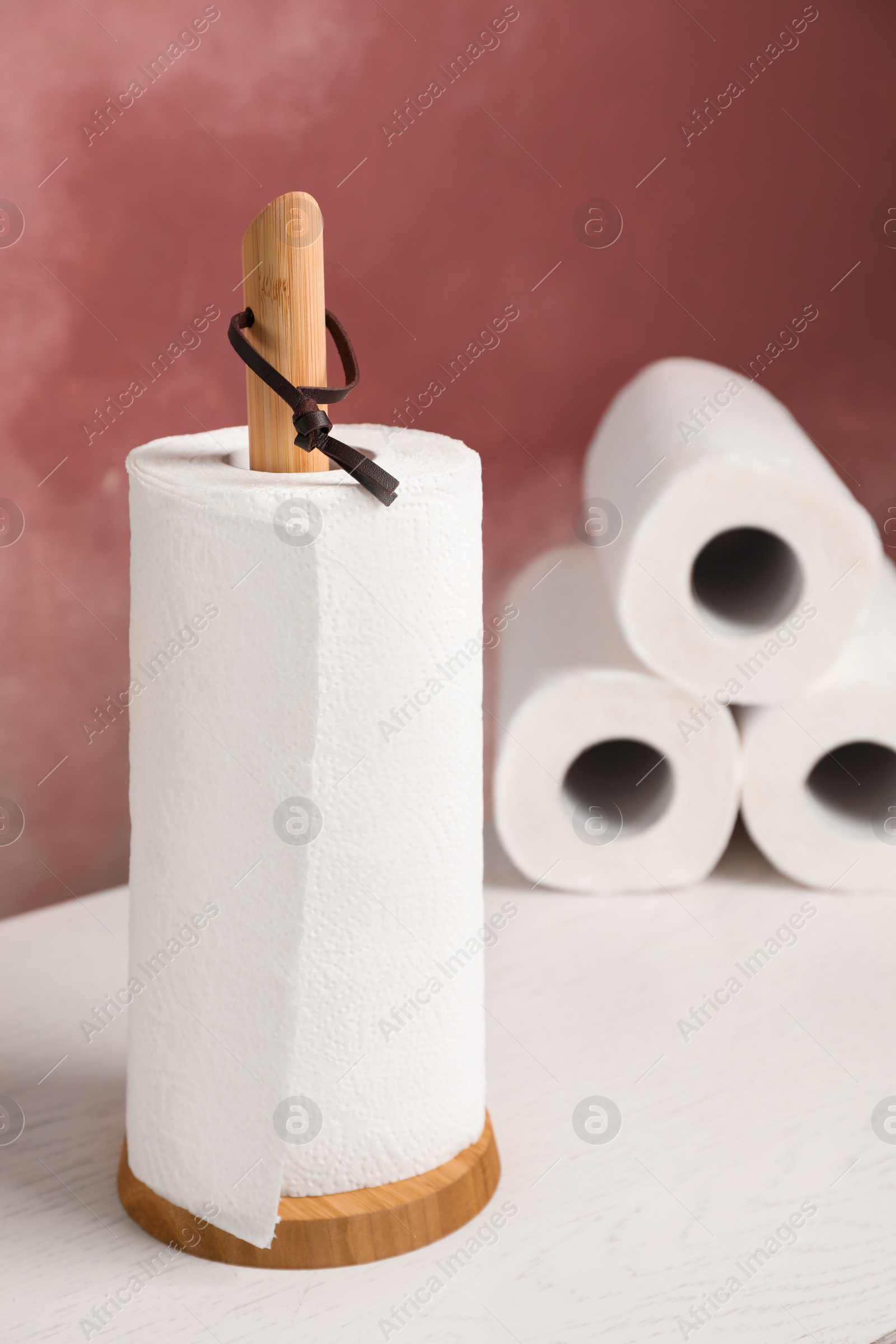 Photo of Holder with roll of paper towels on white wooden table near pink wall