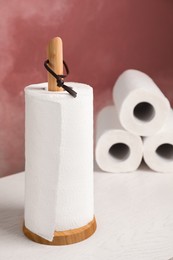 Photo of Holder with roll of paper towels on white wooden table near pink wall
