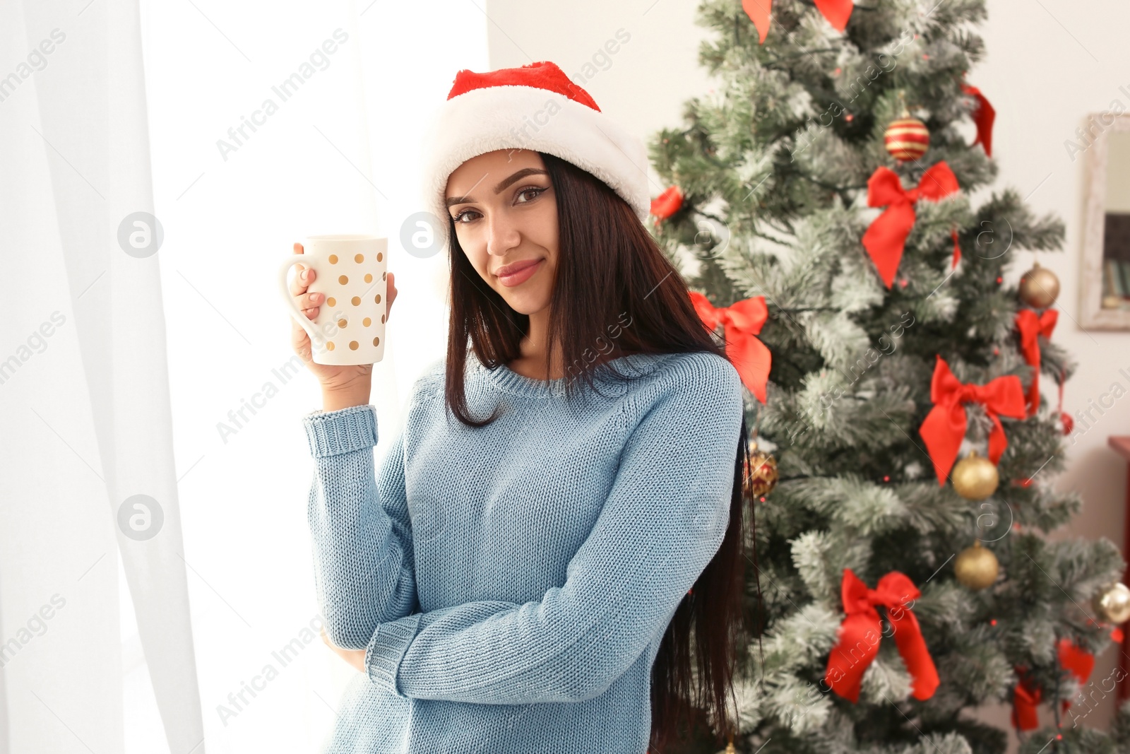 Photo of Beautiful young woman in Santa hat with cup of drink near Christmas tree at home