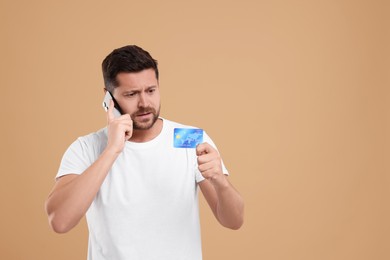 Photo of Upset man with credit card talking on smartphone against beige background, space for text. Be careful - fraud