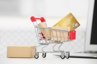 Online payment concept. Small shopping cart with bank card, boxes and laptop on white table, closeup
