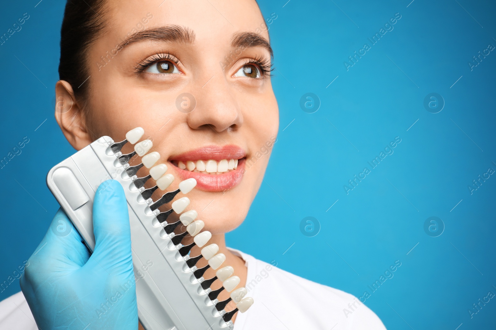 Photo of Doctor checking young woman's teeth color on blue background, closeup. Cosmetic dentistry