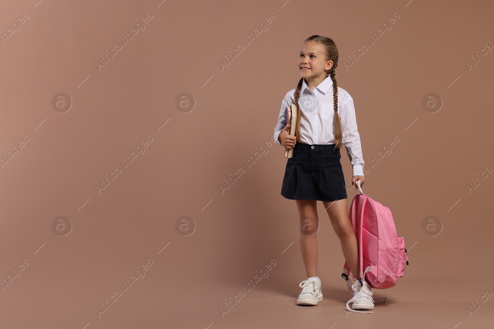 Photo of Happy schoolgirl with backpack and books on brown background, space for text