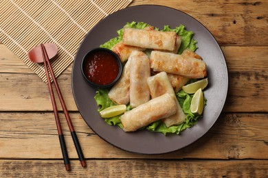 Photo of Plate with tasty fried spring rolls, lettuce, lime, sauce and chopsticks on wooden table, top view