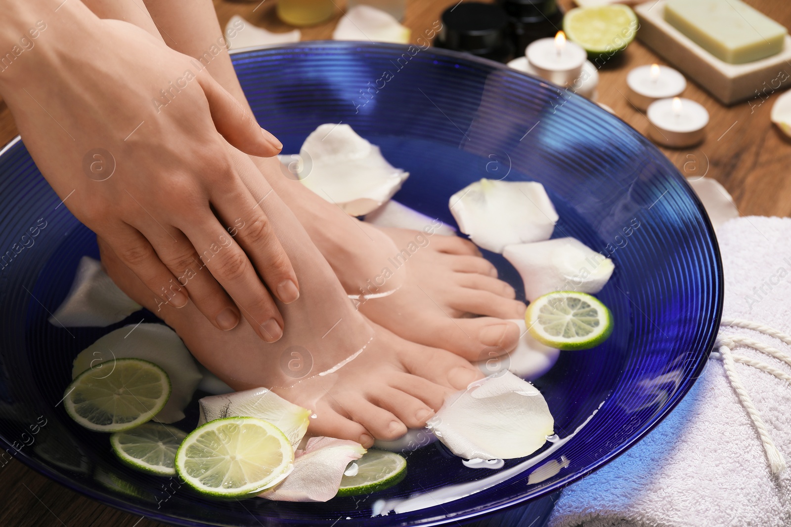 Photo of Woman soaking her feet in bowl with water, petals and lime slices on floor, closeup. Spa treatment