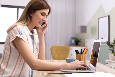 Photo of Young woman talking on phone while working with laptop at desk in home office