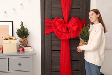 Photo of Woman holding Christmas tree and decorating wooden door with red bow indoors