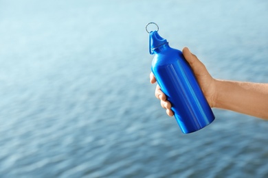 Young sporty man holding water bottle near river on sunny day. Space for text