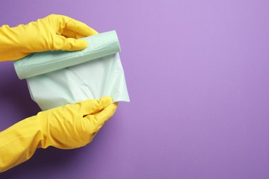 Photo of Janitor in rubber gloves holding roll of color garbage bags over violet background, top view. Space for text
