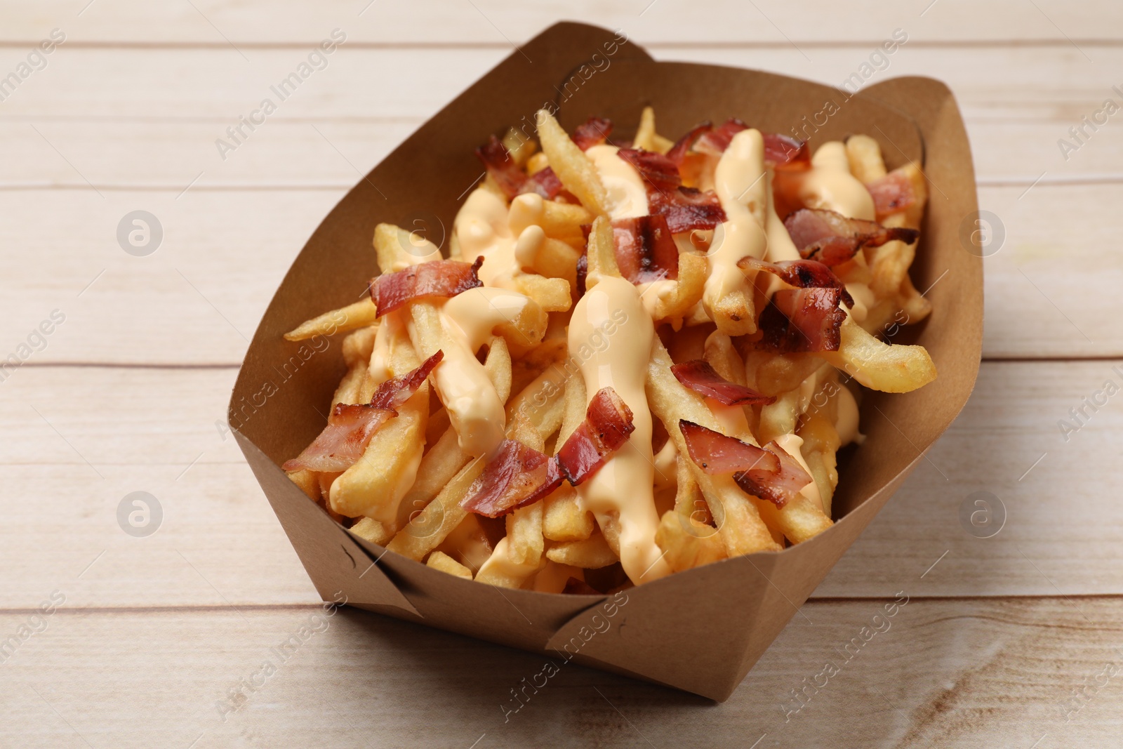 Photo of Tasty potato fries, cheese sauce and bacon in paper container on light wooden table, closeup