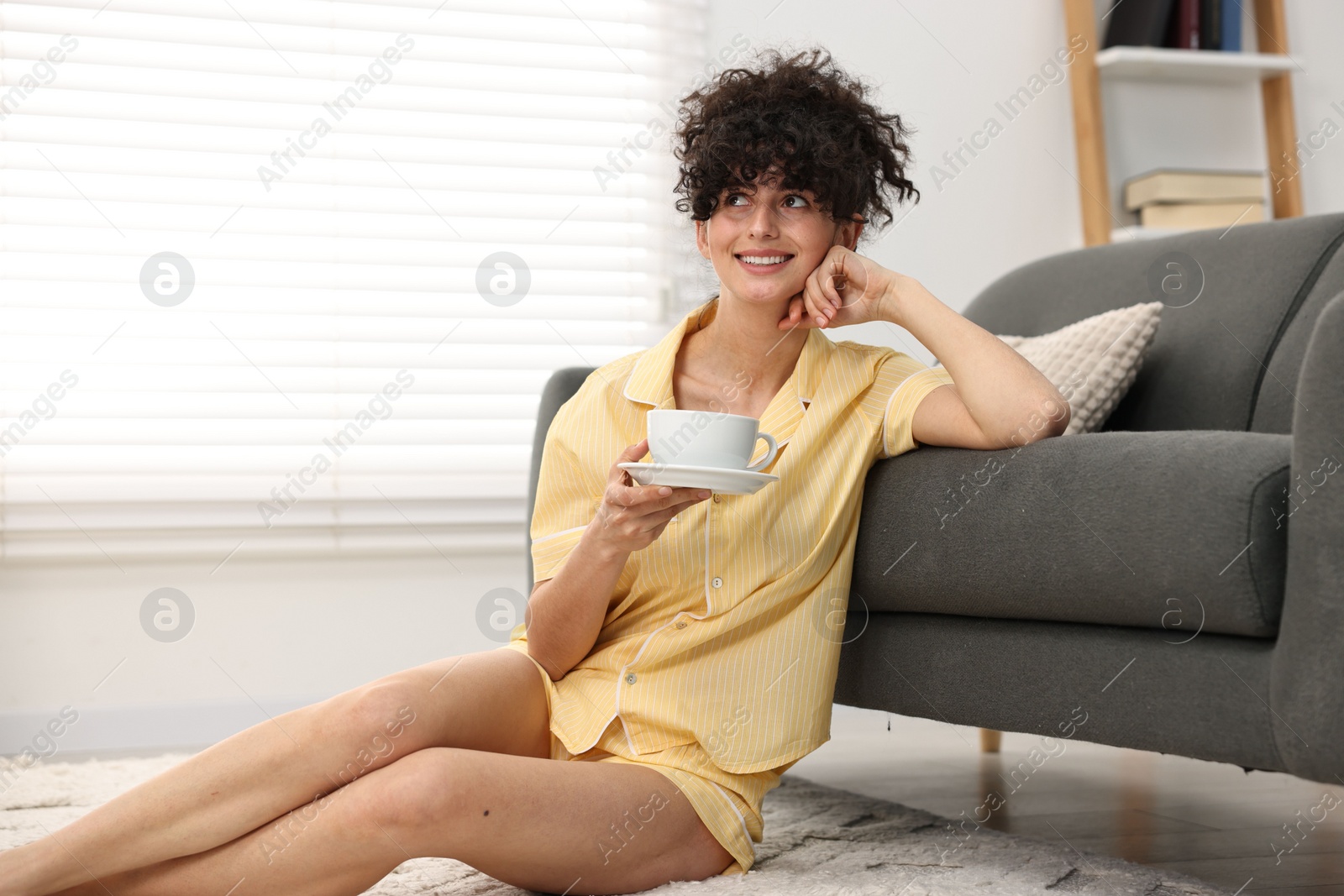 Photo of Beautiful young woman in stylish pyjama with cup of drink on floor at home