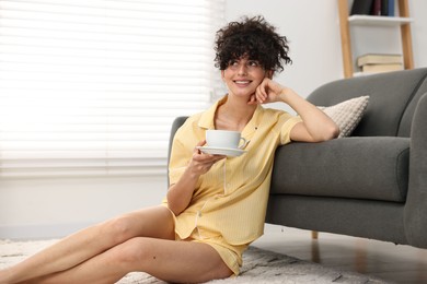 Beautiful young woman in stylish pyjama with cup of drink on floor at home