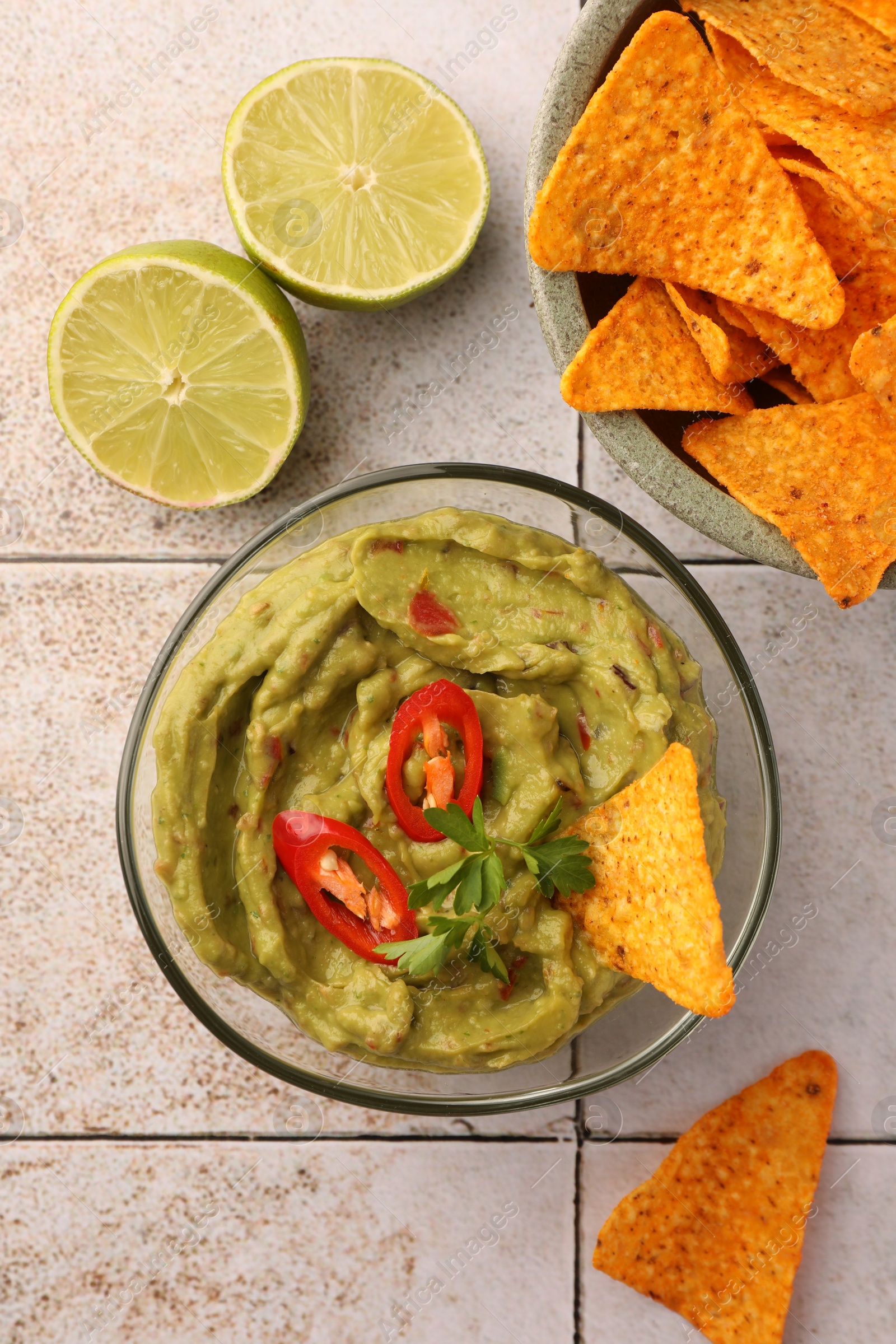 Photo of Bowl of delicious guacamole with chili pepper, nachos chips and lime on white tiled table, flat lay