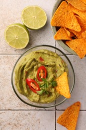 Photo of Bowl of delicious guacamole with chili pepper, nachos chips and lime on white tiled table, flat lay