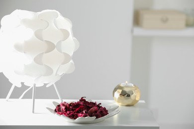 Photo of Plate with aromatic potpourri, lamp and burning candle on white table in room