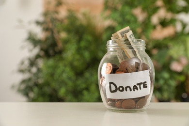 Glass jar with money and label DONATE on table against blurred background. Space for text