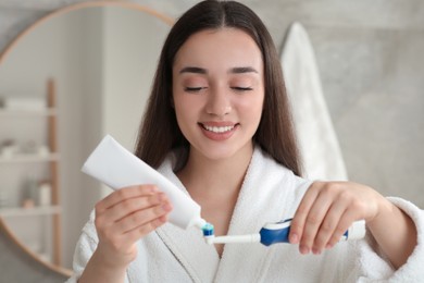 Photo of Young woman squeezing toothpaste from tube onto electric toothbrush in bathroom