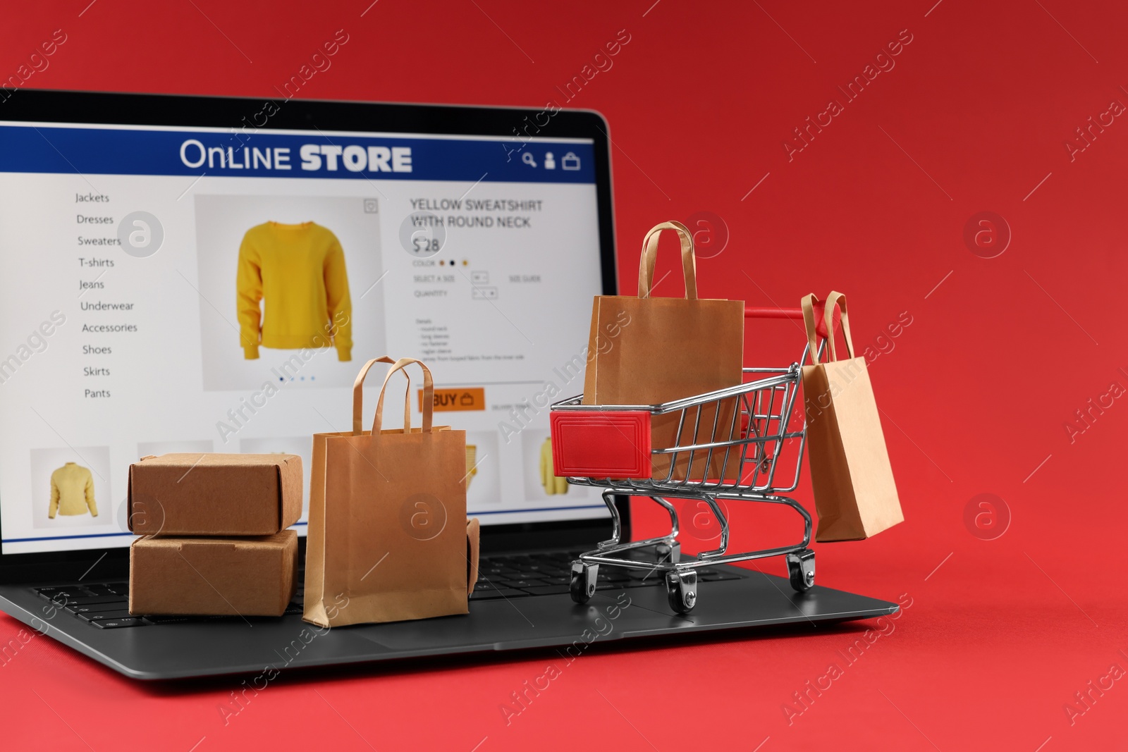 Photo of Online store. Laptop, mini shopping cart and small purchases on red background