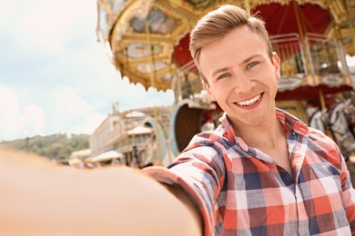 Photo of Young happy man taking selfie near carousel in amusement park