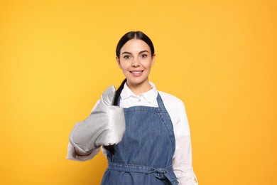 Photo of Young woman in blue jeans apron and oven glove showing thumb up on yellow background