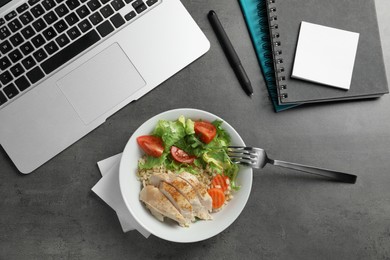 Bowl with tasty food, laptop, fork and notebooks on grey table, flat lay. Business lunch