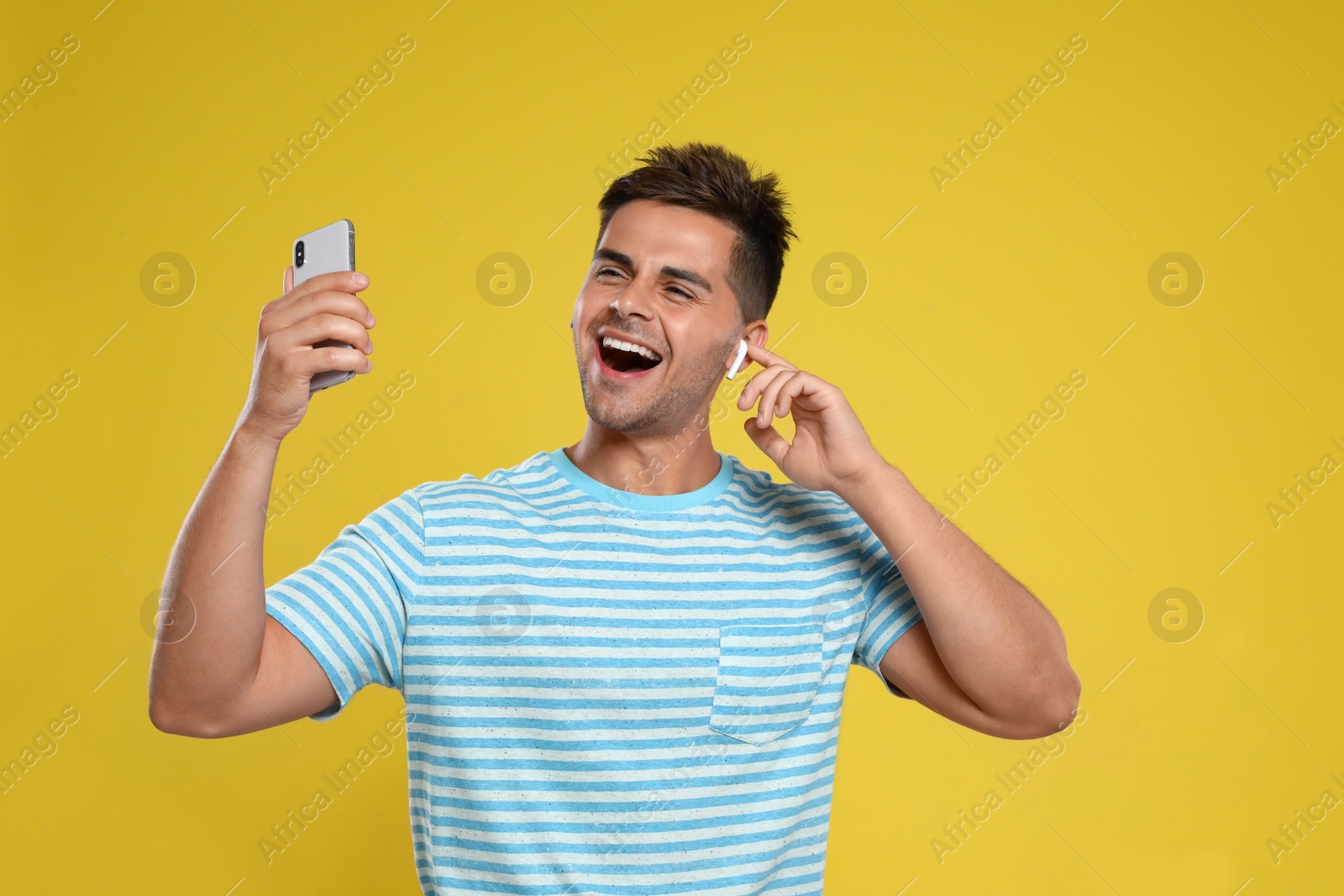 Photo of Happy young man with smartphone listening to music through wireless earphones on yellow background