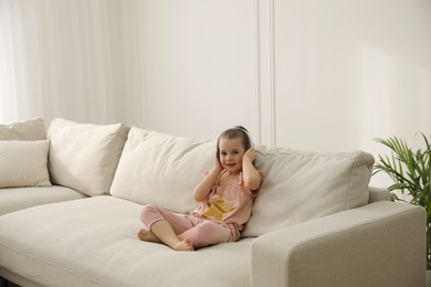 Photo of Little girl with headphones sitting on comfortable sofa in living room