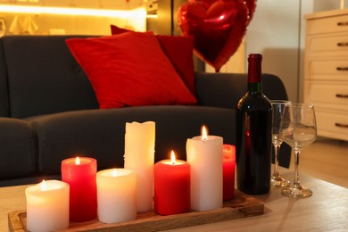 Photo of Romantic atmosphere. Burning candles, bottle of wine and glasses on wooden table near sofa in room decorated for Valentine day