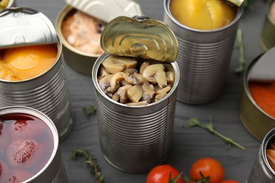 Photo of Open tin cans with mushrooms and different products on gray wooden table, closeup