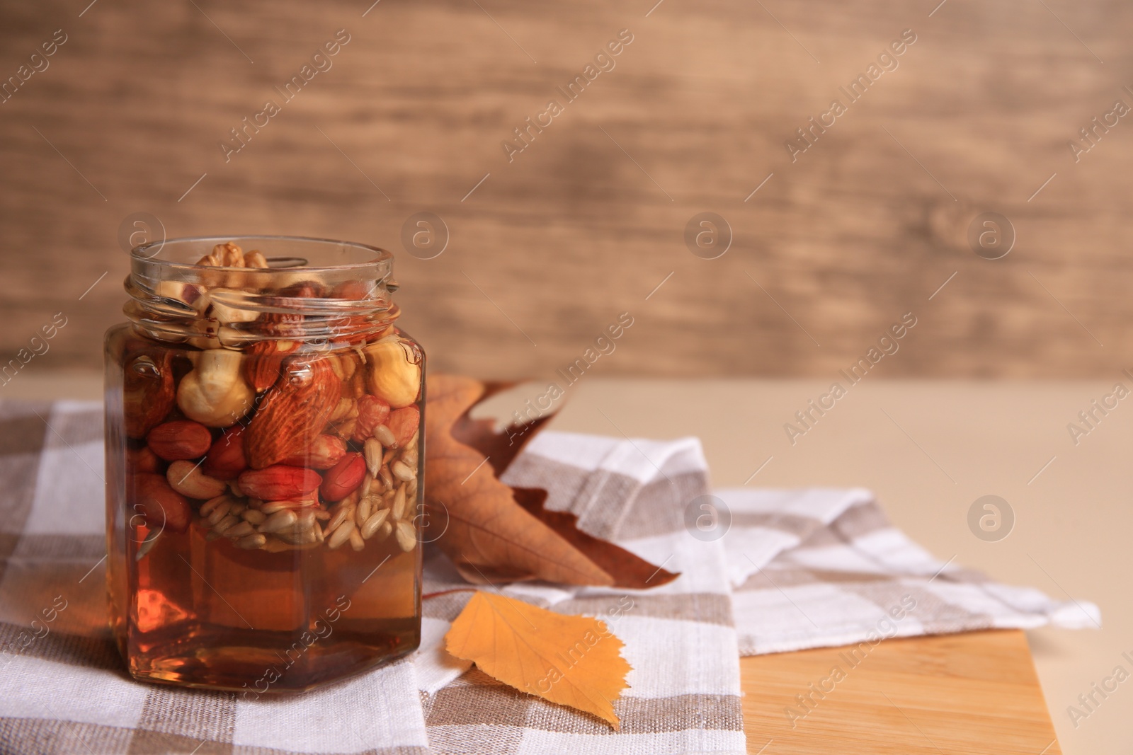 Photo of Different nuts with honey in jar and dry leaves on wooden table. Space for text