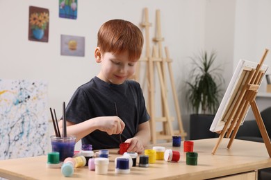 Photo of Little boy painting at table in studio. Using easel to hold canvas
