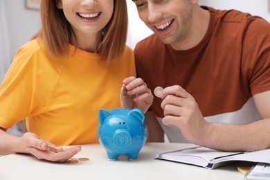 Photo of Couple putting coins into piggy bank at table, closeup. Saving money