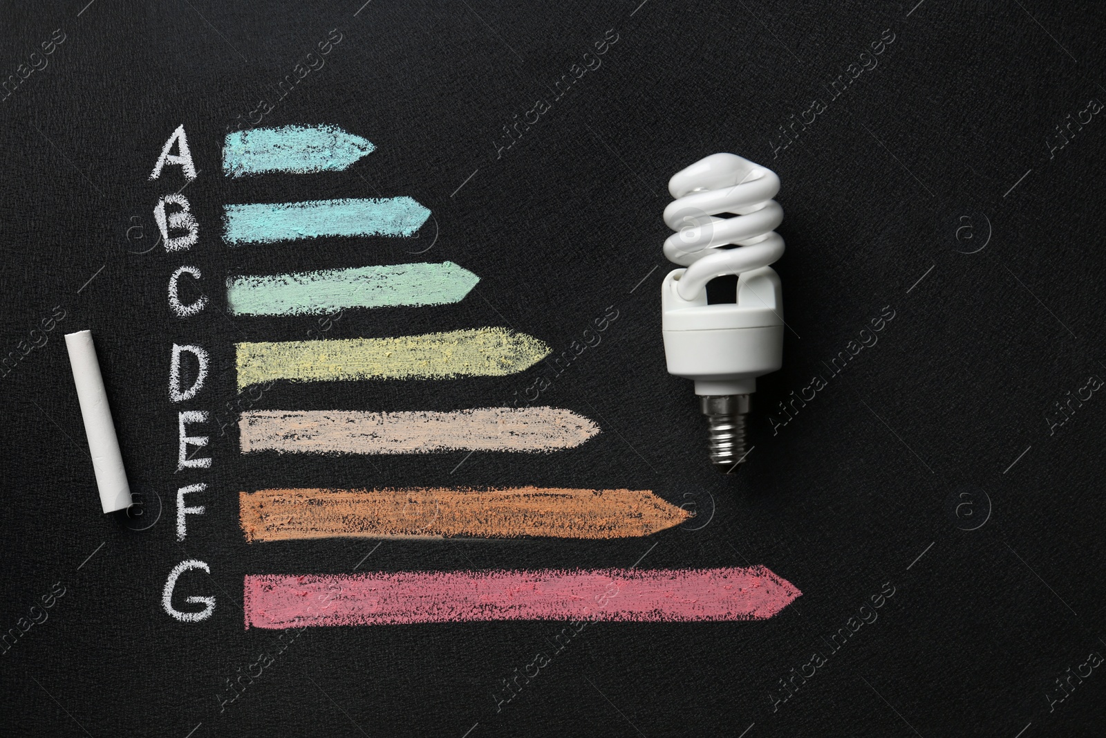 Photo of Energy efficiency rating chart, fluorescent light bulb and chalk on black background, flat lay