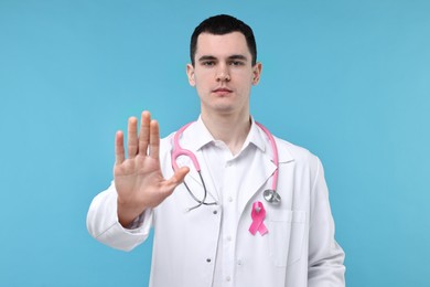 Mammologist with pink ribbon and stethoscope showing stop gesture on light blue background. Breast cancer awareness