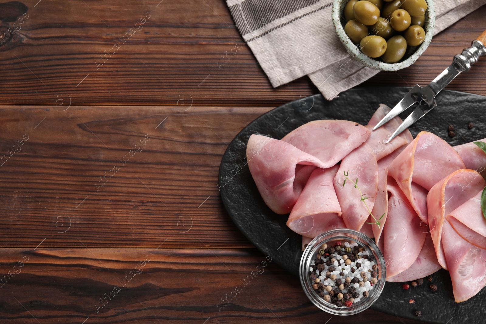 Photo of Tasty ham, olives, sea salt, peppercorns and carving fork on wooden table, flat lay. Space for text