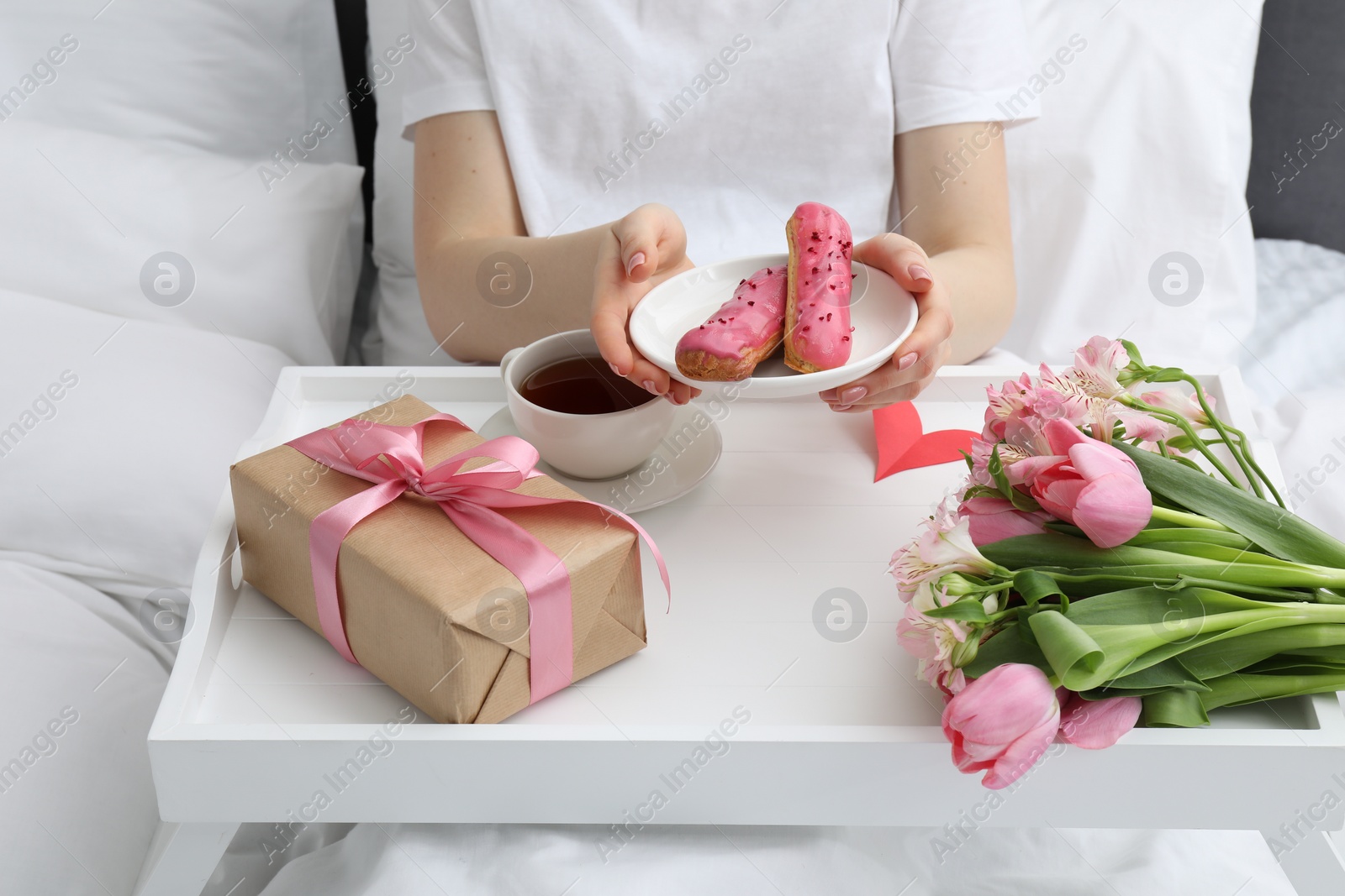 Photo of Tasty breakfast served in bed. Woman with eclairs, tea, gift box and flowers at home, closeup