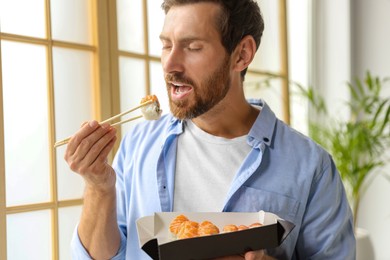 Photo of Man eating tasty sushi rolls and holding box in room, closeup