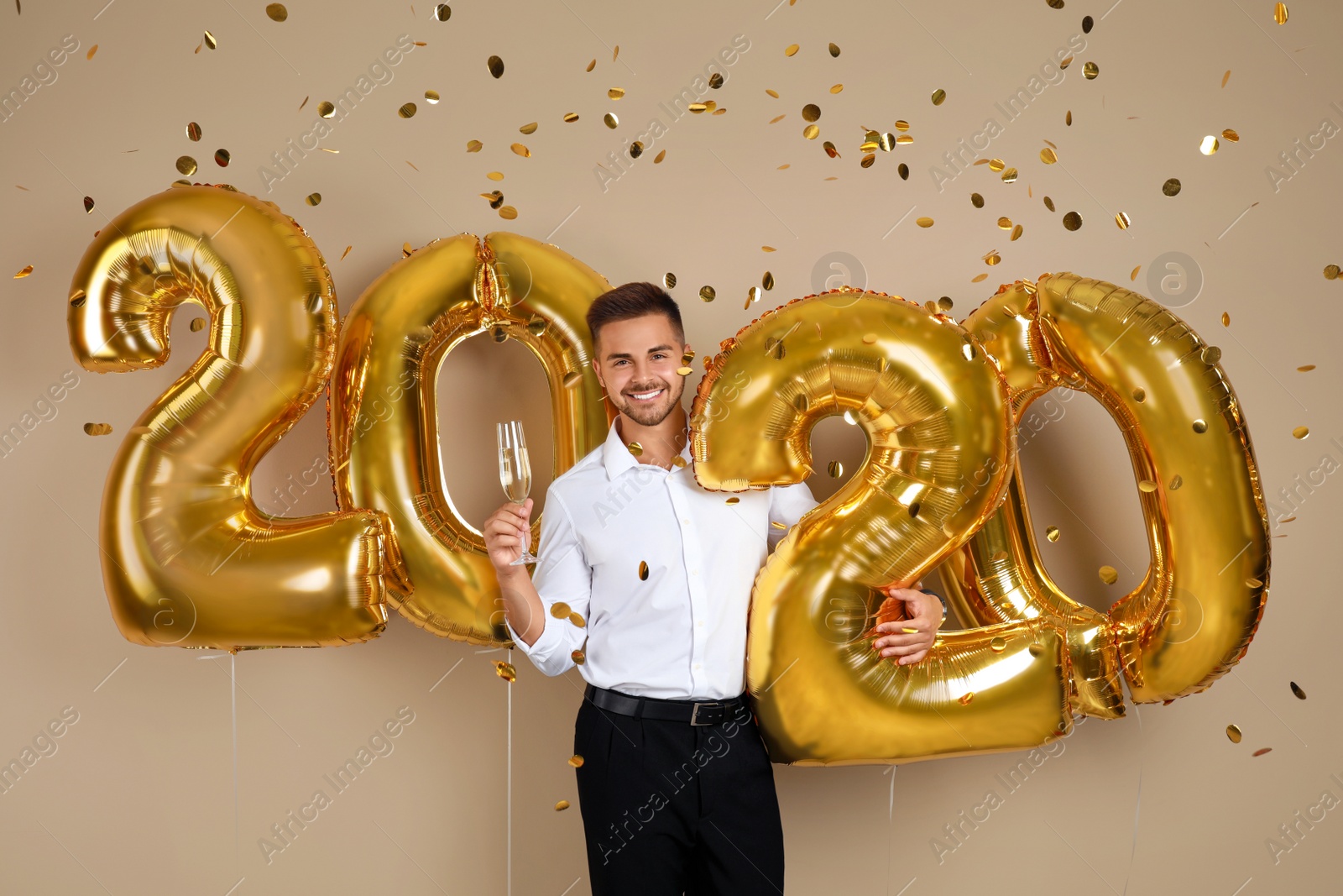 Photo of Happy young man with glass of champagne near golden 2020 balloons on beige background. New Year celebration