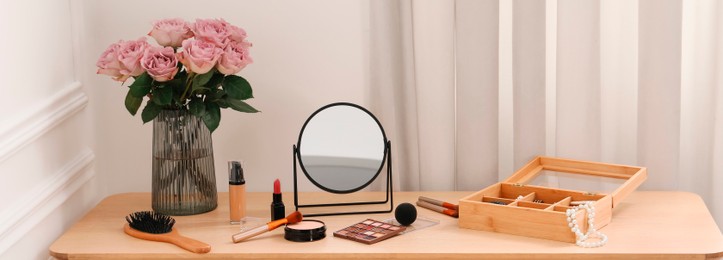 Makeup room. Mirror, cosmetic products, box of jewelry and vase with pink roses on wooden dressing table indoors, banner design