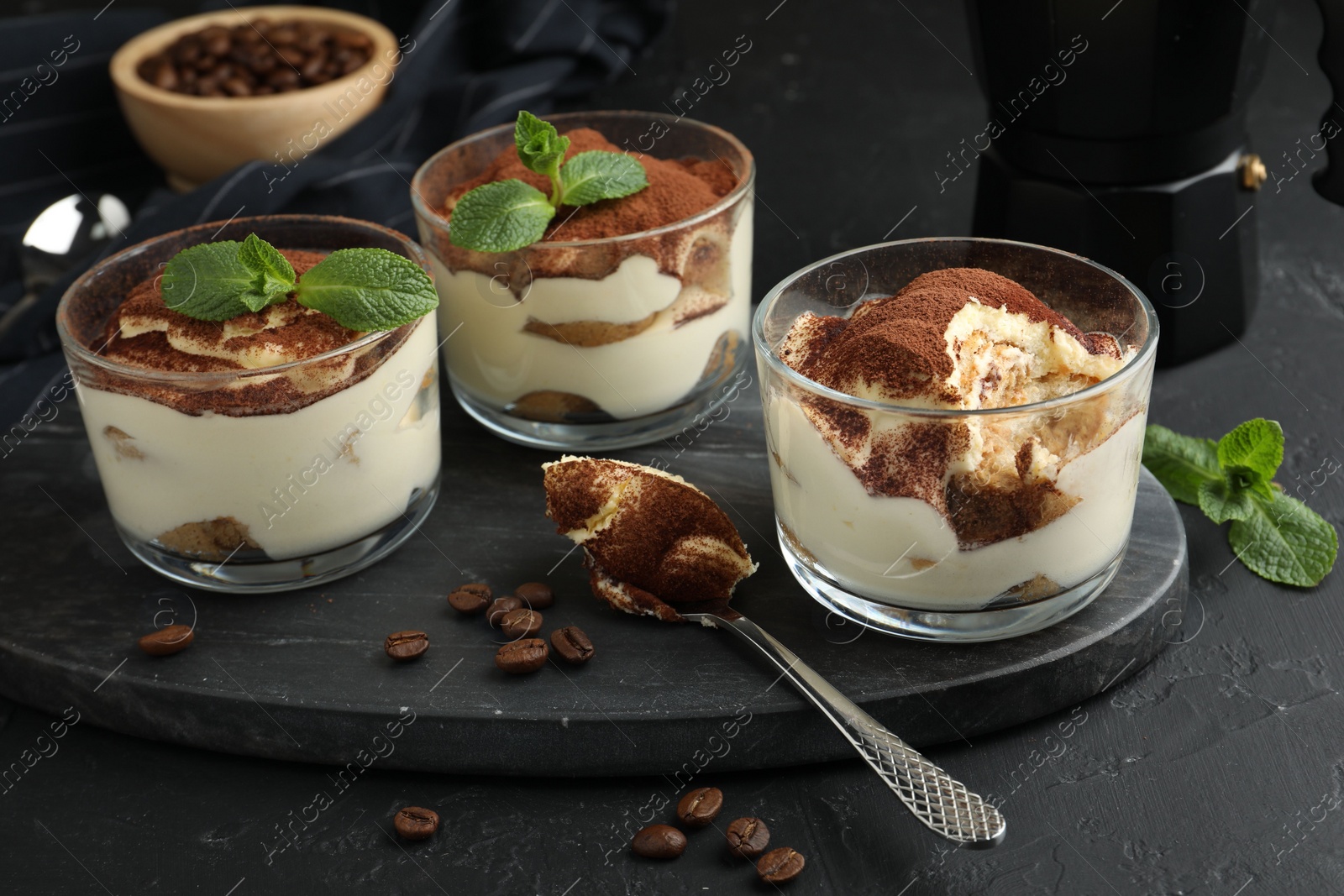 Photo of Delicious tiramisu in glasses, spoon, coffee beans and mint leaves on black table