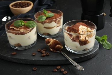 Photo of Delicious tiramisu in glasses, spoon, coffee beans and mint leaves on black table