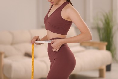 Woman in sportswear measuring waist with tape at home, closeup