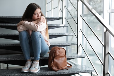 Photo of Upset teenage girl with backpack sitting on stairs indoors. Space for text