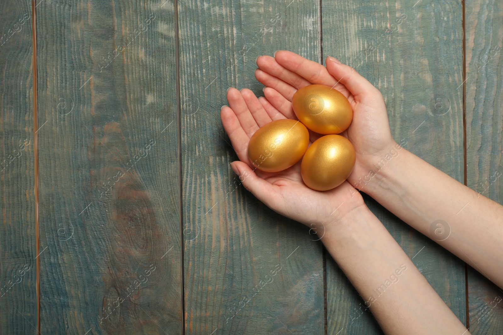 Photo of Woman holding shiny golden eggs at light blue wooden table, top view. Space for text