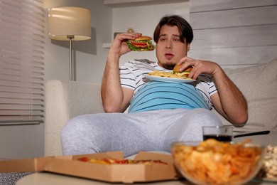 Photo of Overweight man with plate of burgers and French fries on sofa at home