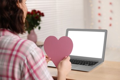 Photo of Valentine's day celebration in long distance relationship. Woman holding pink paper heart while having video chat with her boyfriend via laptop, closeup