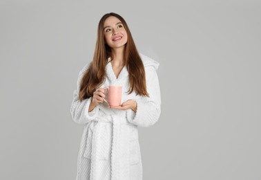 Beautiful young woman in bathrobe with cup of morning drink on light grey background